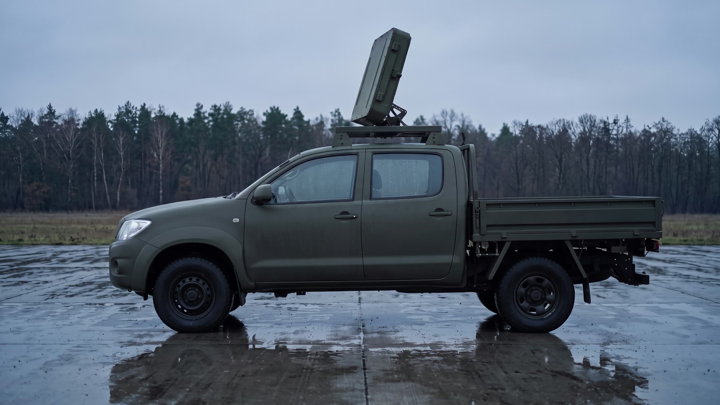 Pickup z radarem na maszcie rozłożonym nad kabiną, mokra płyta postojowa i las w tle
